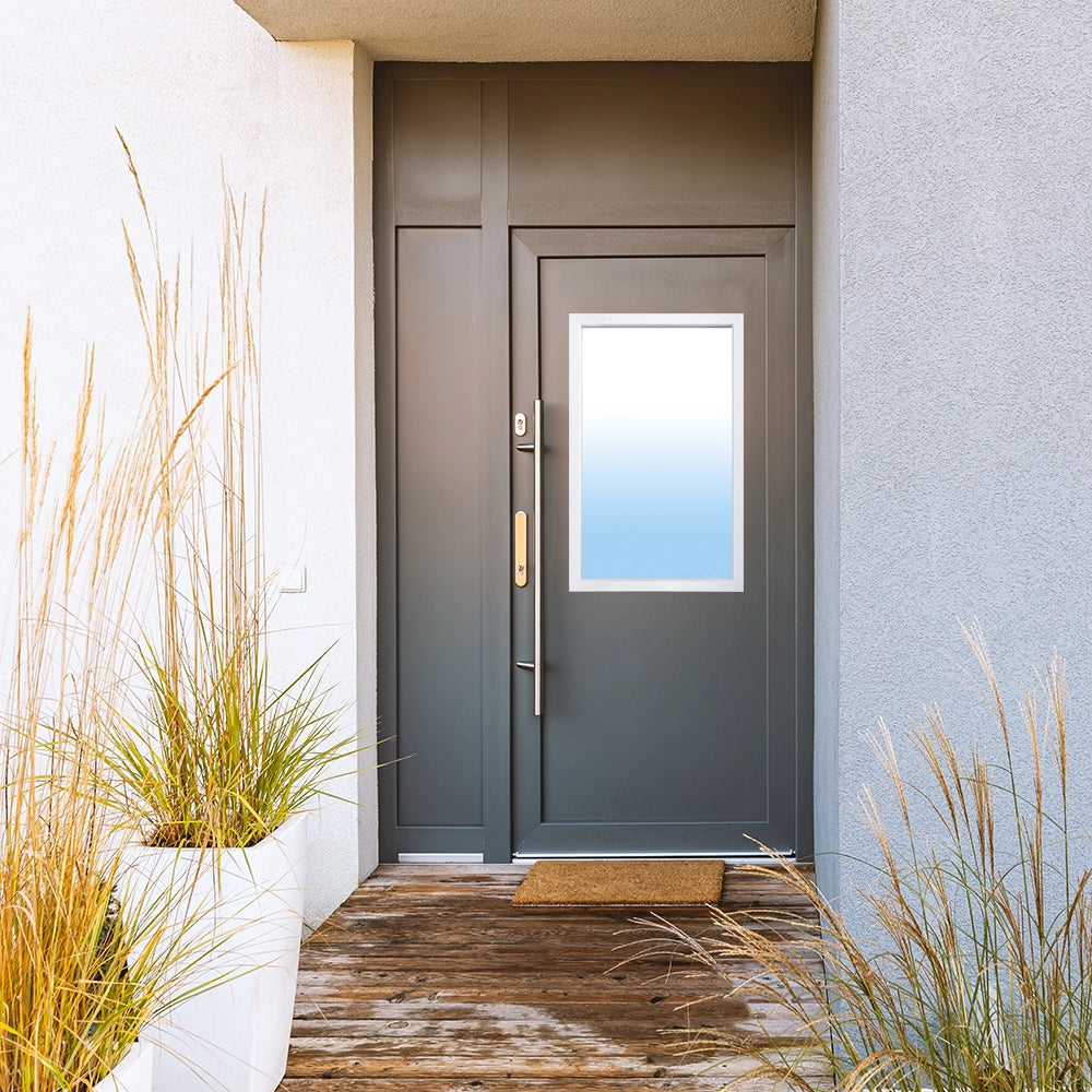A contemporary dark grey front door installed in a modern home's entryway. The door features a white-framed door lite with glass that fades from clear at the top to light blue at the bottom, and is accented with a long, vertical brass handle. The scene includes a wooden deck, a doormat, and large white planters with ornamental grasses on either side of the entrance.