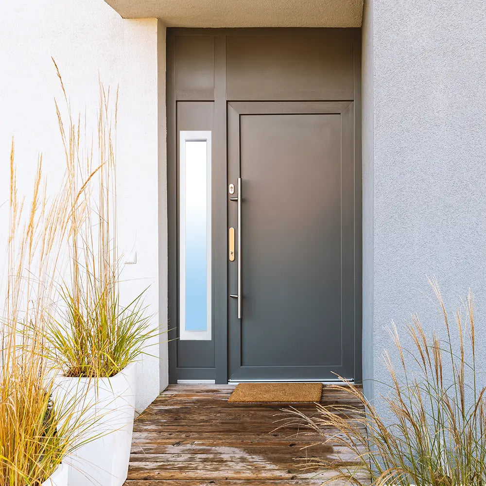 A contemporary home entrance featuring a dark grey door with a long brass handle, a sidelite with a vertical glass window, and modern planters on a wooden porch.